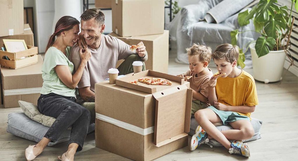 família feliz desempacotando móveis de uma casa nova. do lado esquerdo está um casal de homem e mulher, do lado direito, duas crianças comendo pizza.